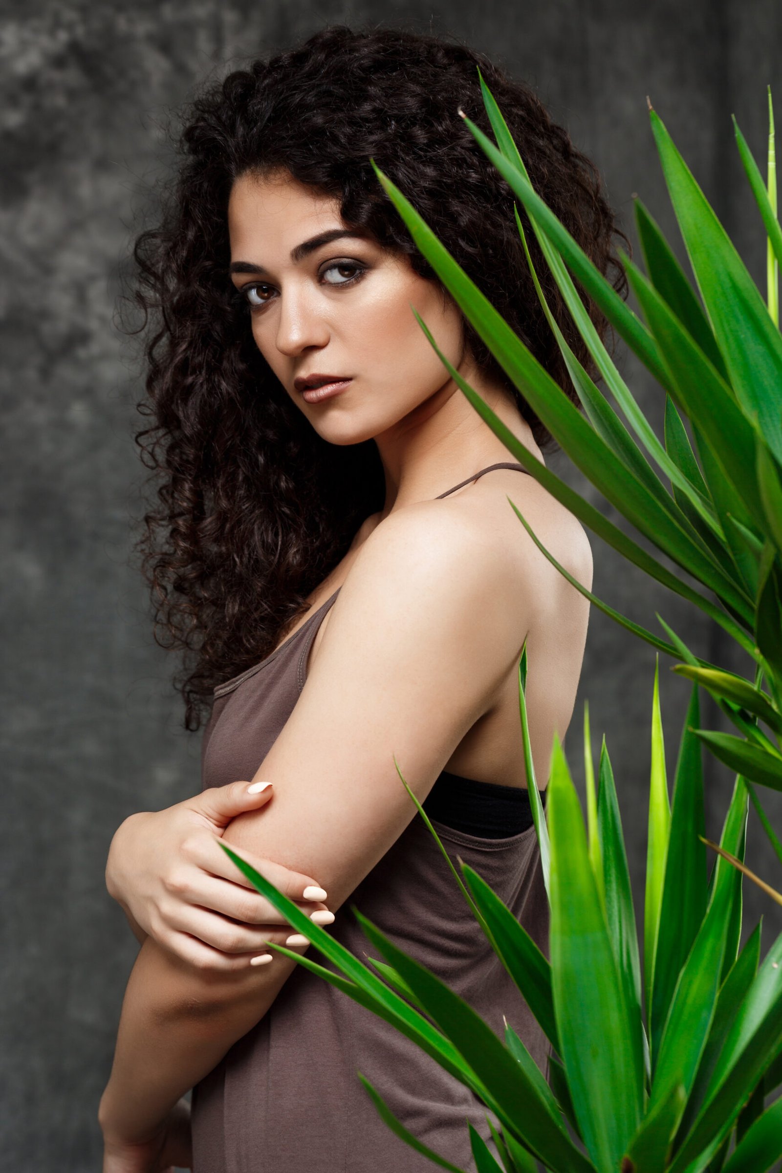 Young beautiful brunette girl in tropical plants looking at camera over grey background.